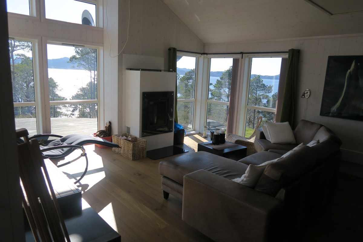 A spacious living area features large windows offering views of the water and surrounding nature. Comfortable seating is arranged around a modern fireplace, with a light wood floor and natural light enhancing the space. A few decorative elements add character to the room.