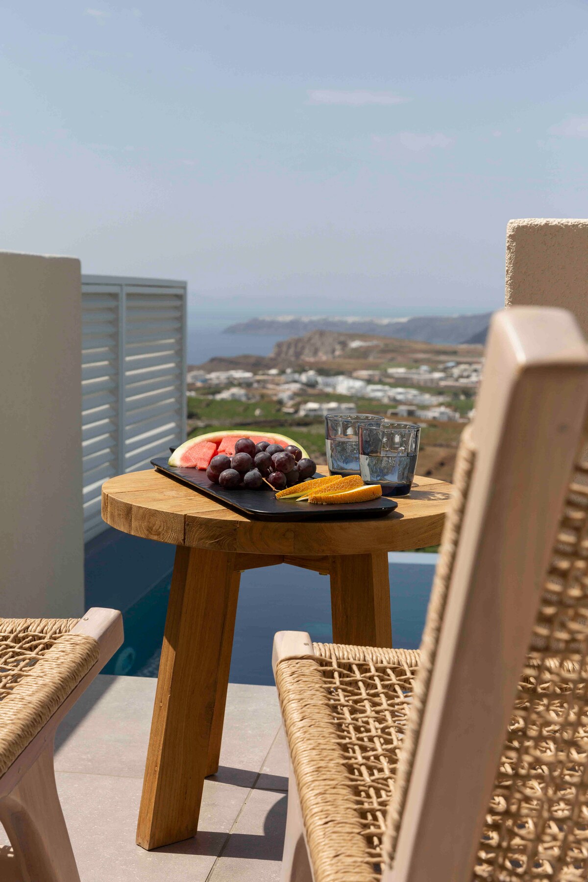 A small wooden table is set with a platter of fresh fruits including slices of watermelon, grapes, and banana, accompanied by two clear glasses. Two woven chairs are positioned alongside the table, with a panoramic view of the sea and landscape visible in the background.