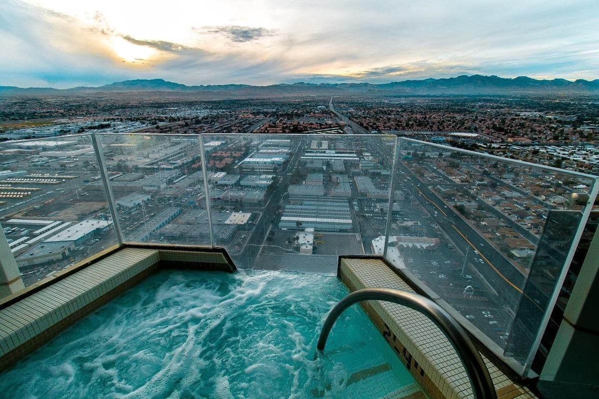 The image captures a luxurious outdoor Jacuzzi overlooking the vast landscape of Las Vegas. The horizon features distant mountains under a softly lit sky, with the city sprawling below. A clear glass railing provides unobstructed views of the surroundings.