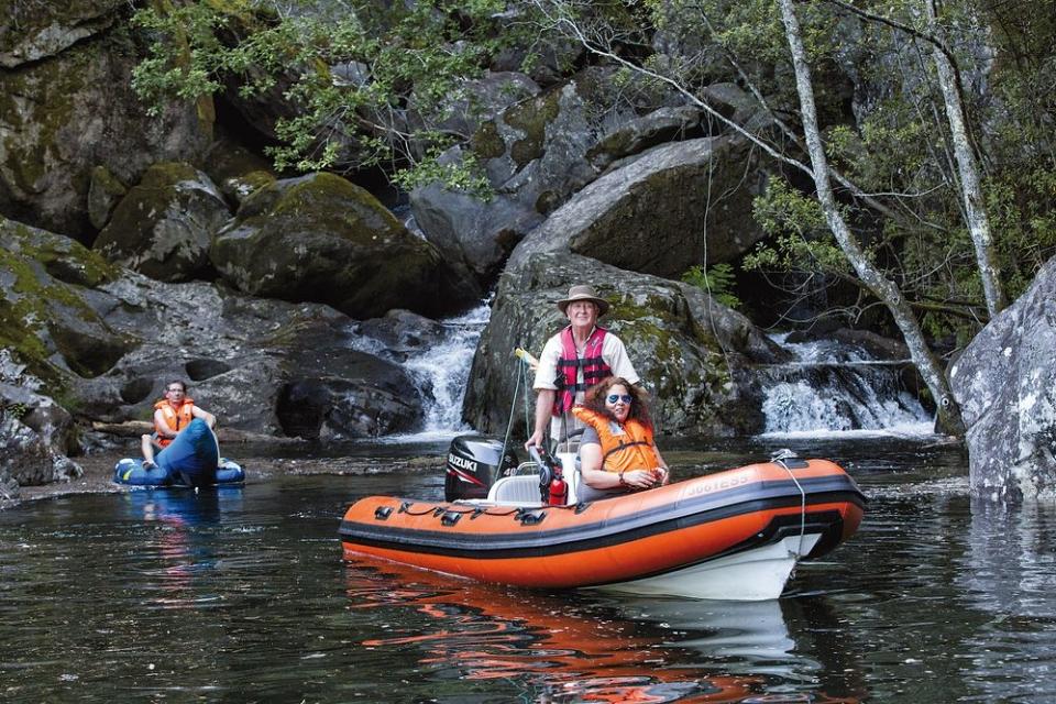 An inflatable boat is anchored in calm waters, featuring individuals wearing life jackets. Lush greenery and rugged rocks surround the scene. A small waterfall can be seen in the background, enhancing the natural setting, while another person is seen in a kayak nearby.