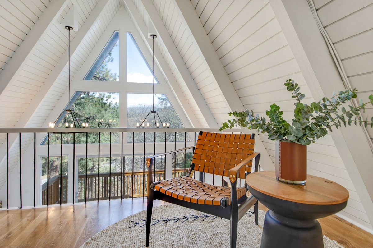 A loft area features a woven chair beside a round side table. Natural light fills the space through a large triangular window, offering views of the surrounding trees. A decorative plant sits on the table, adding a touch of greenery to the modern design.