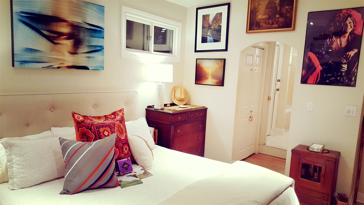 A cozy bedroom features a king-size bed with decorative pillows, nestled against a headboard. Art pieces adorn the walls, complementing a warm ambience. A bedside table holds small items, while a wooden dresser adds character. A doorway leads to the bathroom, providing easy access.