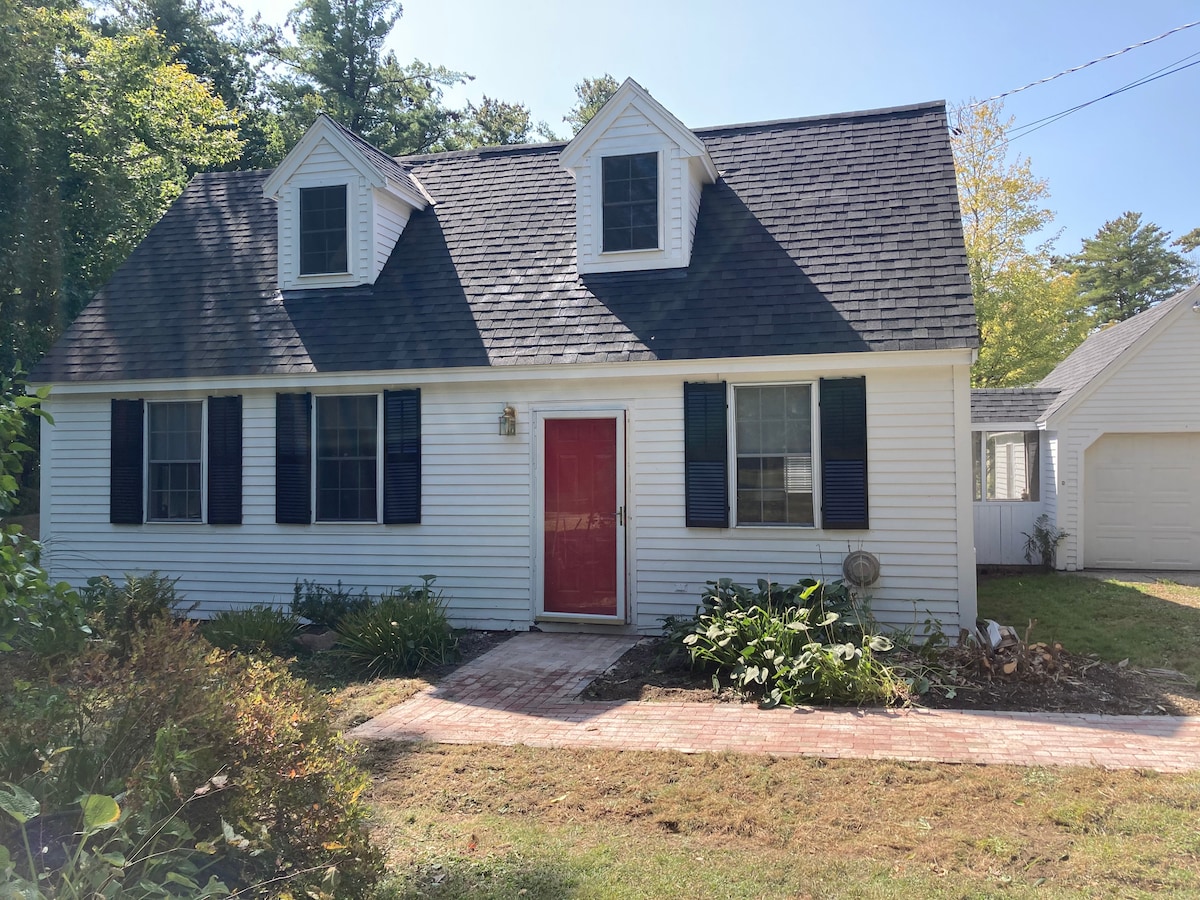 The cottage features a Cape-style exterior with white siding and dark shutters. A red front door draws the eye, while the landscaped area includes shrubs and small trees. Natural light highlights the windows, and a brick pathway leads to the entrance.