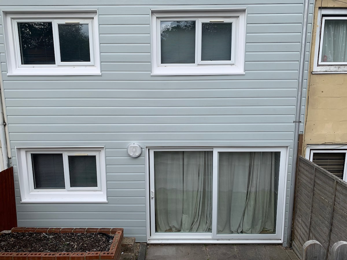 The exterior of a two-story building is shown, featuring pale blue siding and multiple windows. A large glass sliding door, partially covered by sheer curtains, serves as the main entrance on the ground floor. A small gravel area with a decorative planter is visible in the foreground.