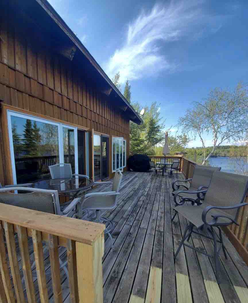 A spacious wooden deck extends along the house, surrounded by tall trees. Several chairs and a table create a comfortable outdoor seating area. Large windows reflect natural light and offer views of the serene lake nearby.