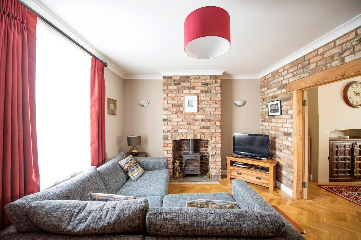 An inviting living area features a spacious sectional sofa facing a brick fireplace. Large windows are adorned with red curtains, allowing natural light to fill the space. A flat-screen television is positioned on a wooden stand, complementing the cozy atmosphere.