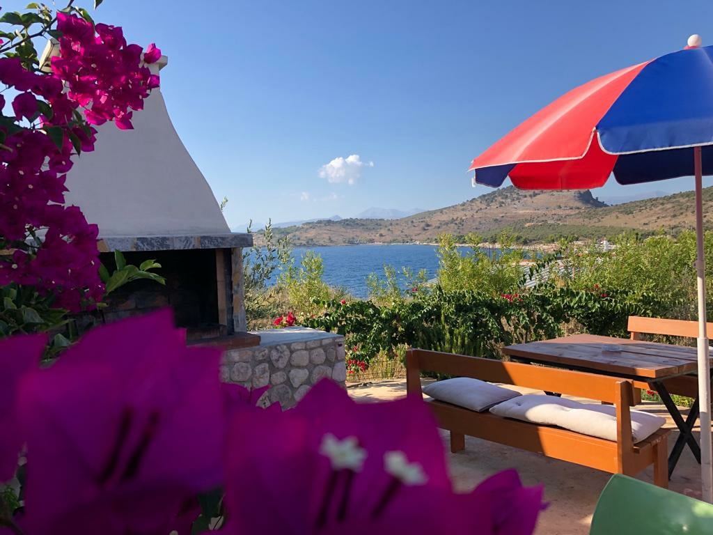 A vibrant outdoor space features a stone barbecue surrounded by lush greenery and blooming flowers. A wooden table and seating area are present under a large, colorful umbrella, offering views of the tranquil water beyond.