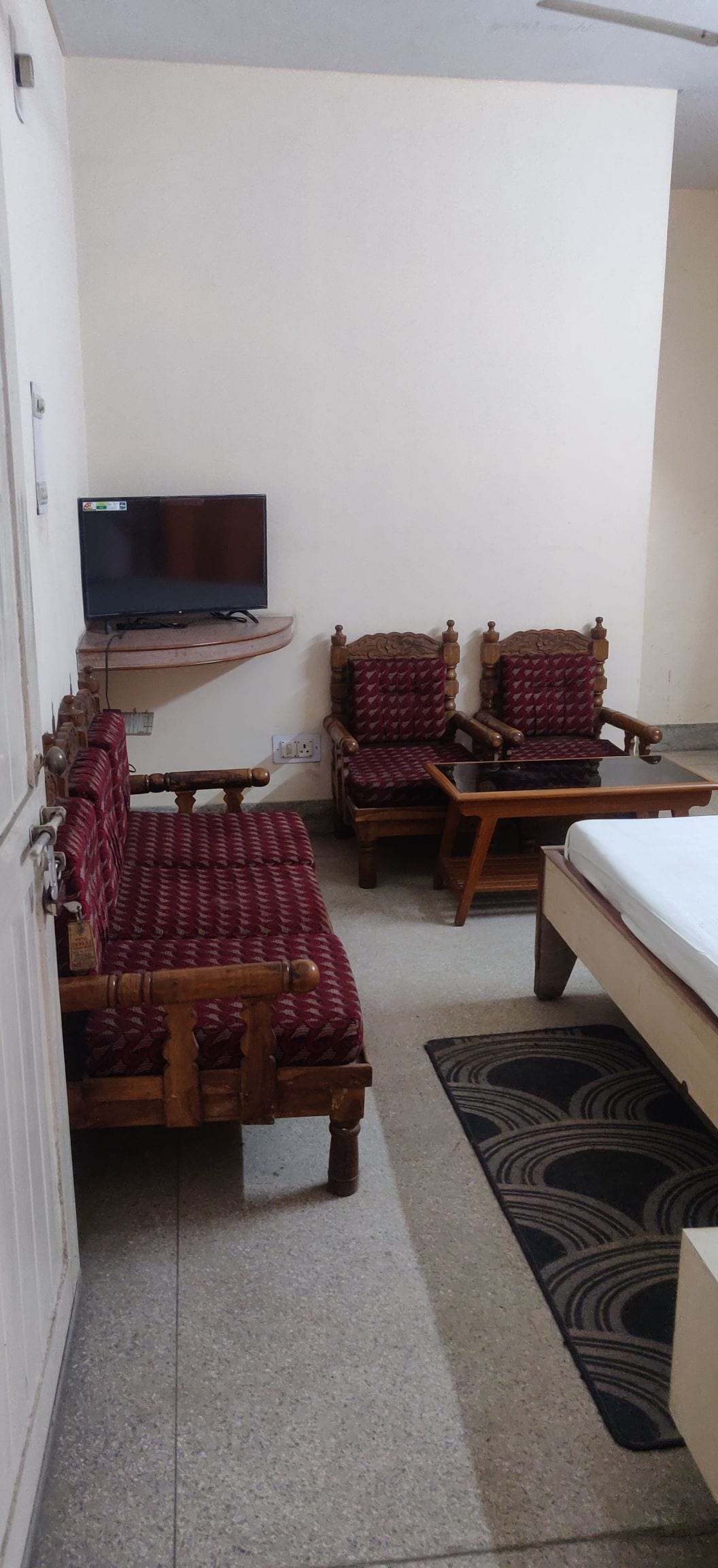 A cozy room features a spacious bed beside a patterned rug. Two traditional wooden chairs with cushions are positioned next to a small wooden table. A television is mounted on the wall, providing entertainment options in a comfortably relaxed environment.