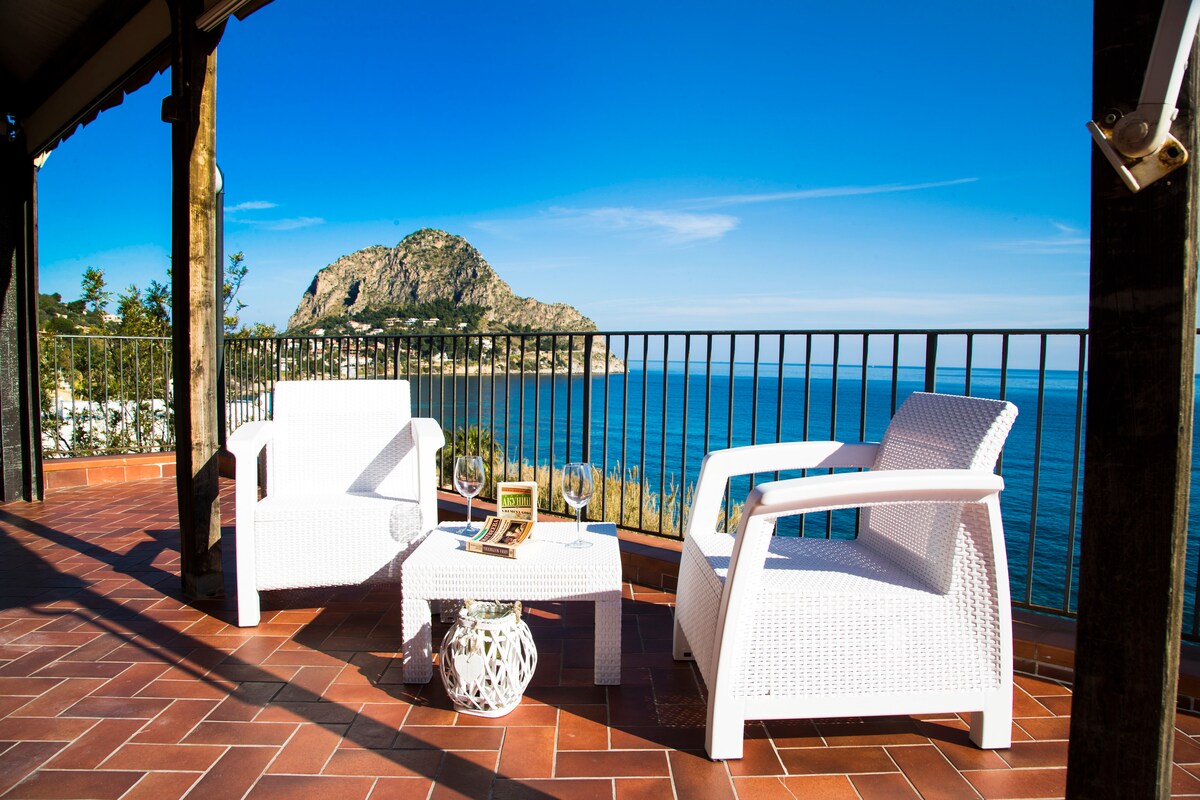 A private terrace features two white armchairs and a small coffee table, providing a serene setting for outdoor relaxation. The captivating view encompasses the azure sea and a distant rocky landscape under a clear blue sky.