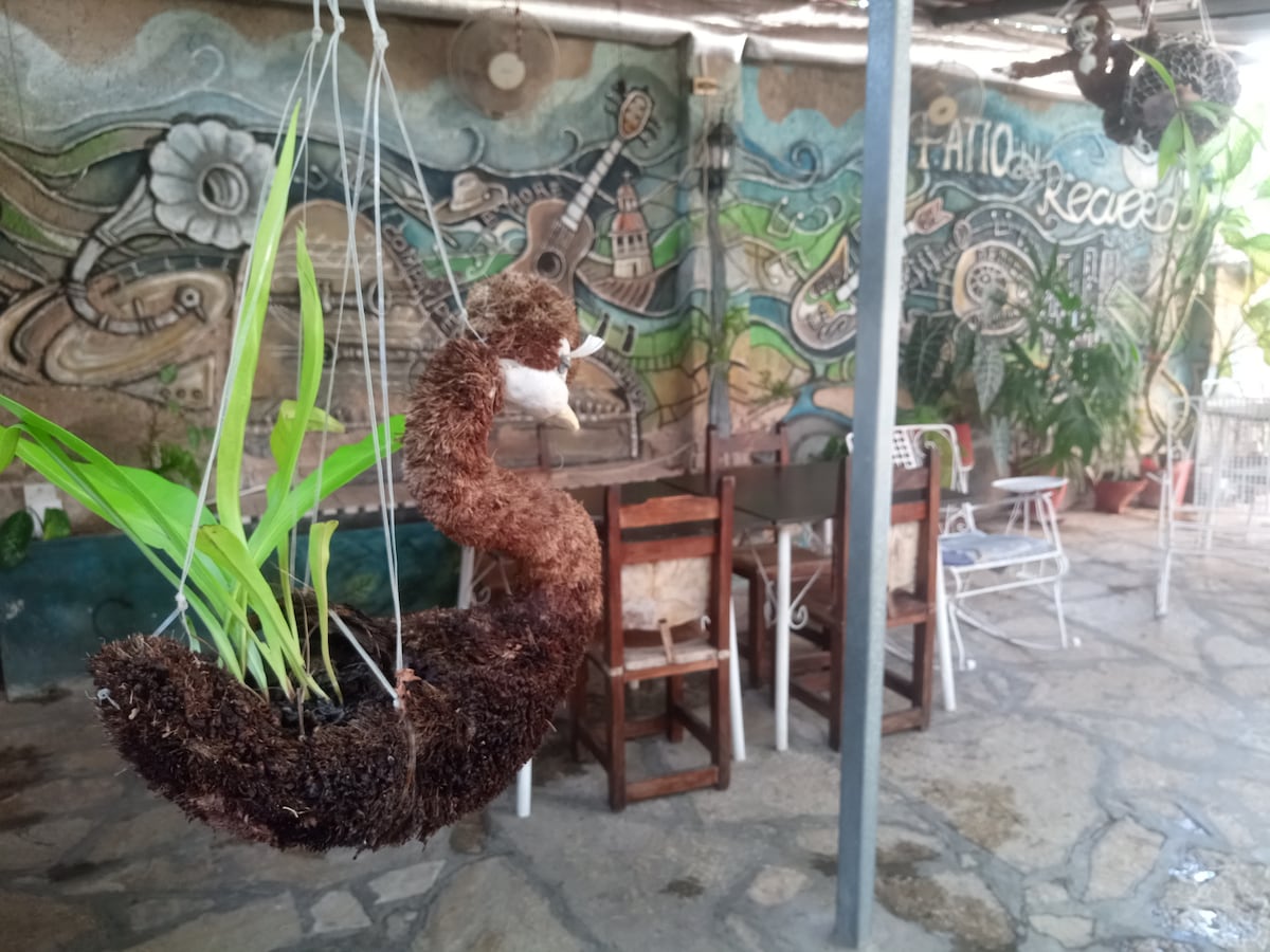 A cozy patio area is shown, featuring a hanging planter crafted from natural materials. The background displays a colorful mural, while wooden chairs are arranged around a stone floor, creating an inviting space for guests to relax.