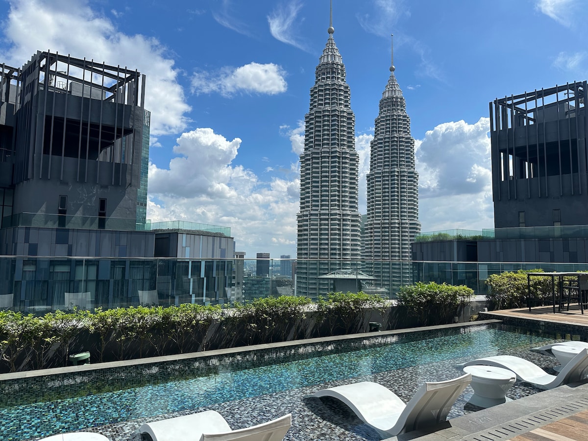 The rooftop pool area features lounge chairs arranged beside a shimmering water surface. Towering in the background are the iconic twin towers of KLCC, framed by a clear blue sky dotted with clouds.