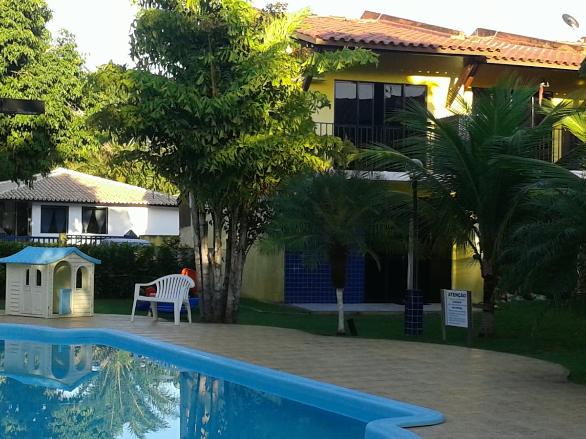 A swimming pool reflects the surrounding greenery and a two-story yellow house is visible in the background, displaying large windows. A white plastic chair is positioned near the pool, and a small playhouse is set in the grassy area beside the water.