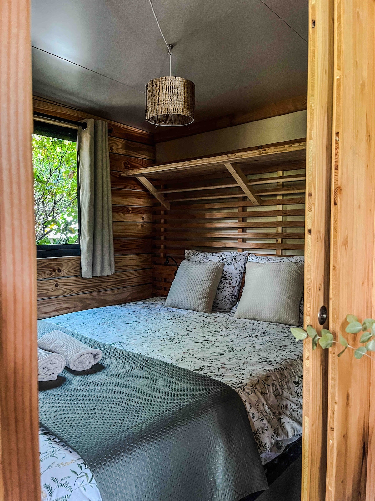 A cozy sleeping area features natural wood paneling and a spacious bed dressed in a floral pattern. Soft linens are neatly arranged, with two towels placed at the foot of the bed. A window allows for natural light and views of greenery outside.