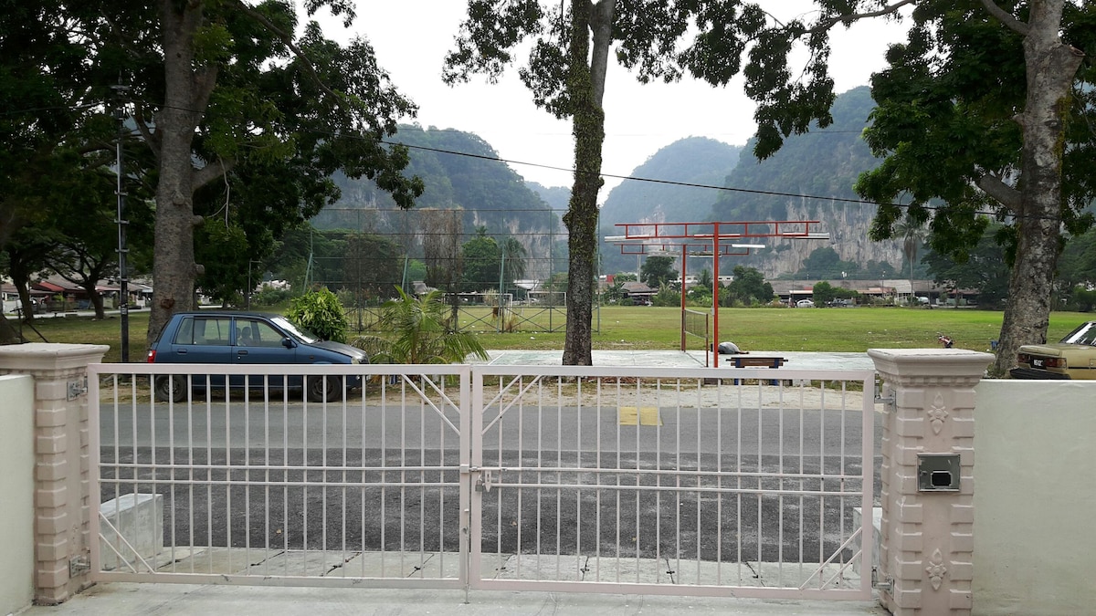 A gated entrance frames a view of a grassy area and distant hills. Lush trees line the edges of the scene, while a few cars are parked along the street. The sky is overcast, creating a muted atmosphere.