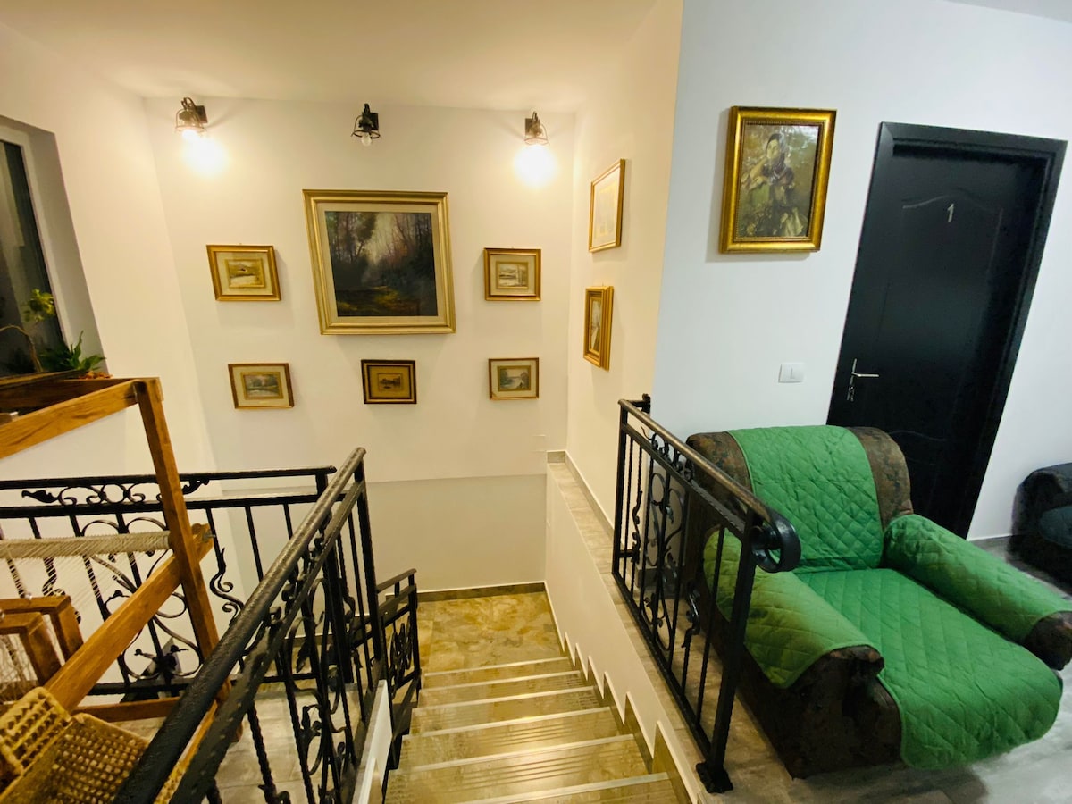 A staircase is framed by elegant metal railings, leading to an upper floor. The walls feature an arrangement of framed artwork, while soft lighting highlights the vibrant colors. A green upholstered armchair is positioned nearby, creating a welcoming space for relaxation.