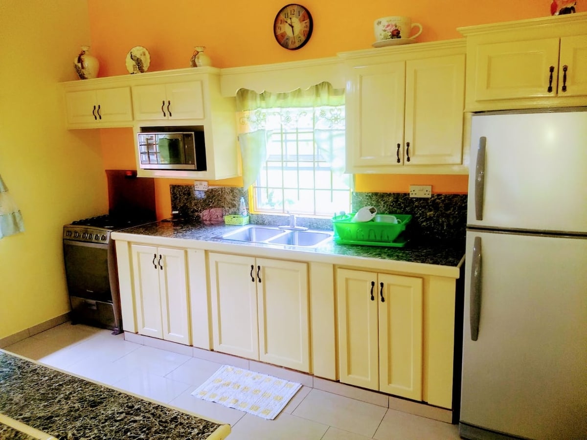 A kitchen features yellow cabinetry and a dark granite countertop. A double sink is positioned underneath a window, allowing natural light to illuminate the space. The kitchen includes an oven, microwave, and a refrigerator, with a decorative clock and dish rack adding a homey touch.