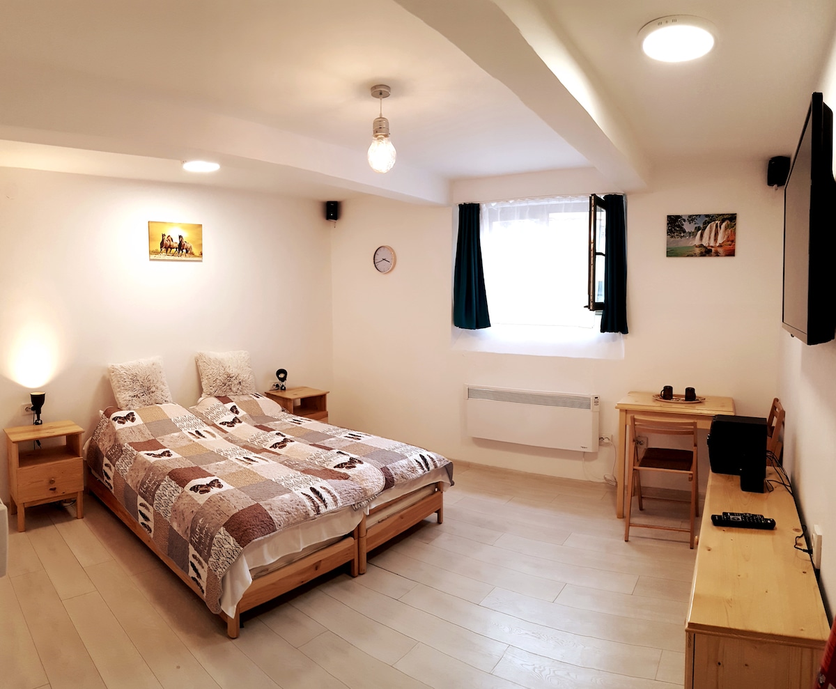 A cozy studio layout features a spacious bed dressed in a patterned quilt. A wooden desk and chair are positioned near a window, allowing natural light. Wall art and a clock add character to the bright, minimalistic decor.