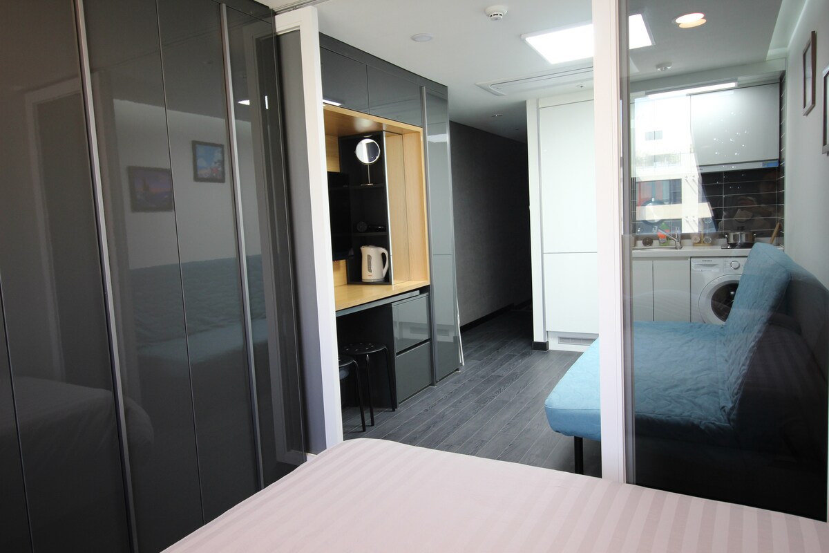 A modern living space is shown, featuring sleek cabinetry and a comfortable seating area. A kitchenette is visible, equipped with essential appliances. Soft natural light enters through a window, while the floor is covered with dark, smooth material, enhancing the room's contemporary feel.