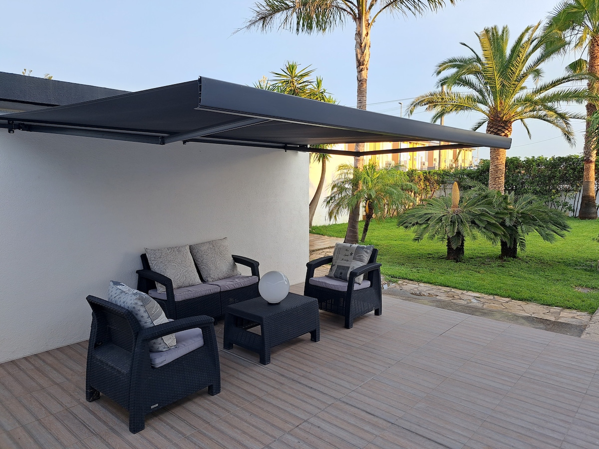 A shaded outdoor seating area features a comfortable arrangement of black wicker chairs and a matching table. Soft cushions add comfort, while palm trees and lush greenery create a serene backdrop. The patio surface is tiled, promoting a clean, inviting space for relaxation.