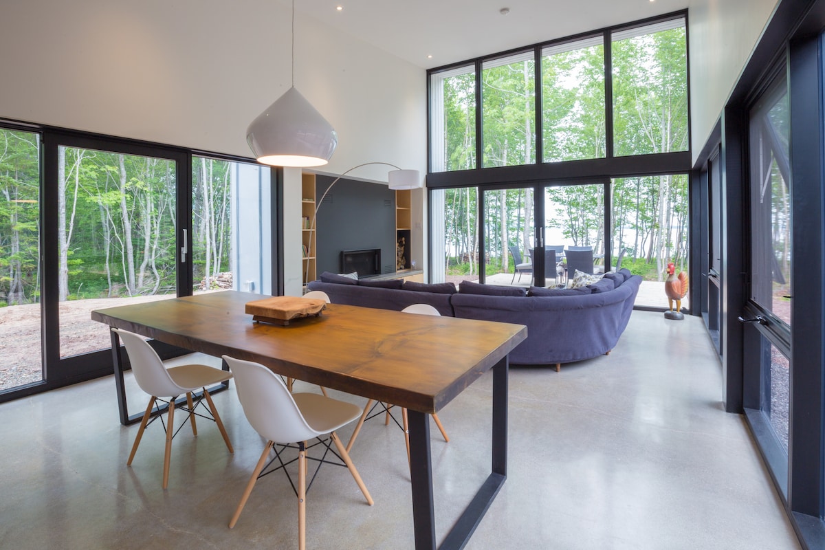 An open concept living space features a wooden dining table with white chairs adjoining a cozy lounge area. Large glass windows provide a view of the surrounding forest, while a sectional couch invites relaxation. Natural light brightens the airy ambiance.