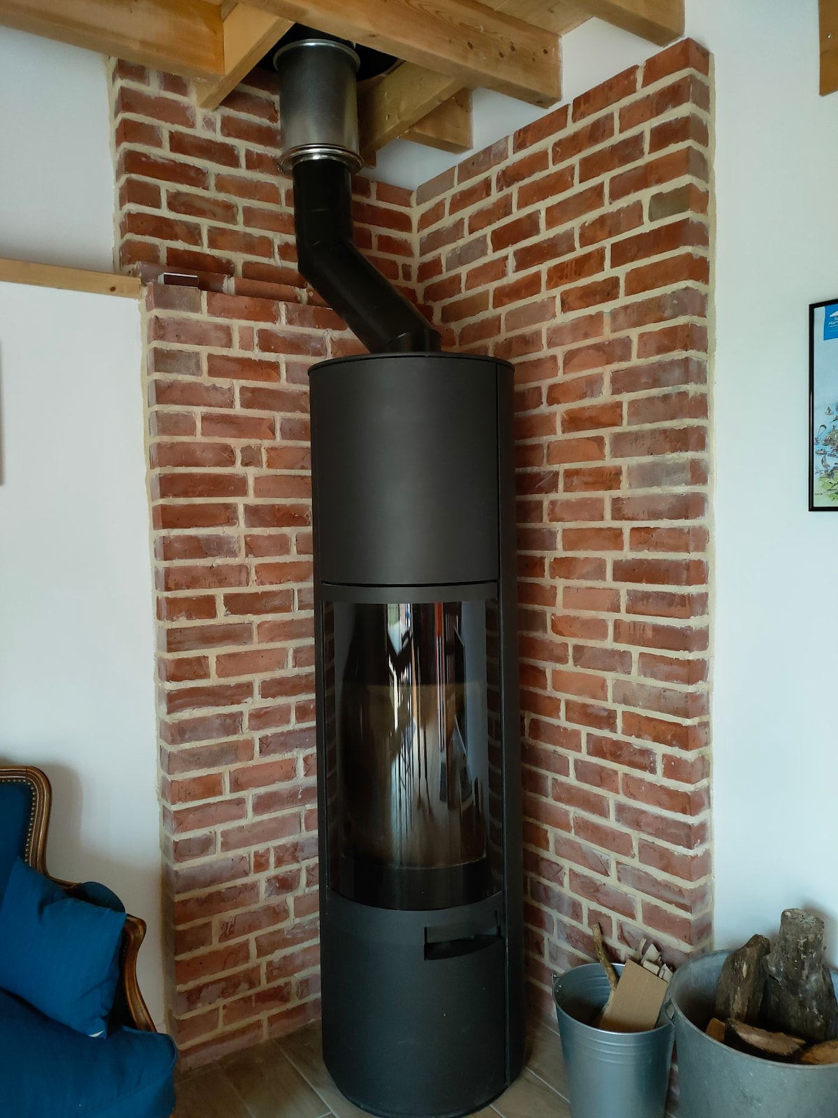 A modern wood stove is positioned against a brick wall, with glass panels showcasing the fire within. The design features a cylindrical shape and a sleek black finish, providing warmth to the living space. A metal bucket and firewood are placed nearby for convenience.