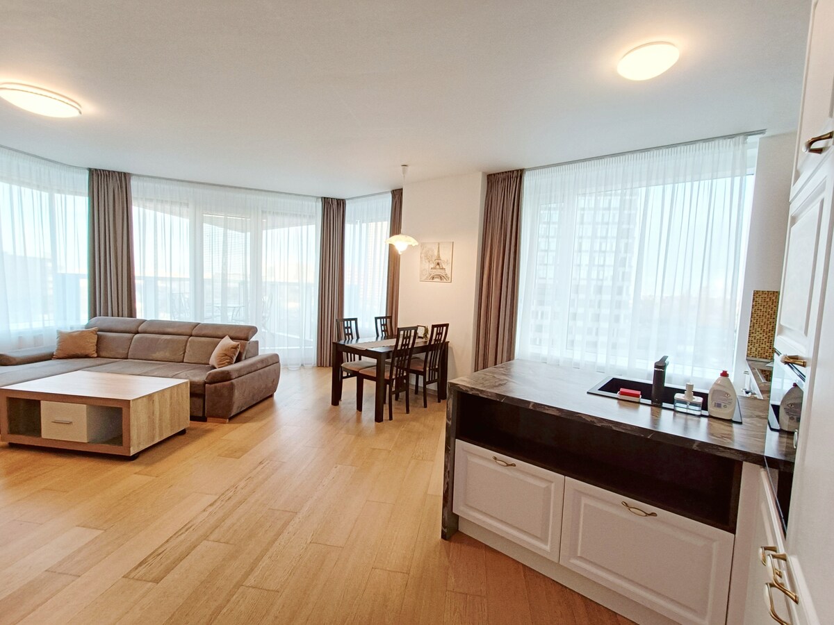 A spacious living area features large windows with sheer curtains, allowing natural light to fill the space. A comfortable sectional sofa and a wooden dining table with six chairs are present. A sleek kitchen counter with minimal clutter is visible, enhancing the open layout.