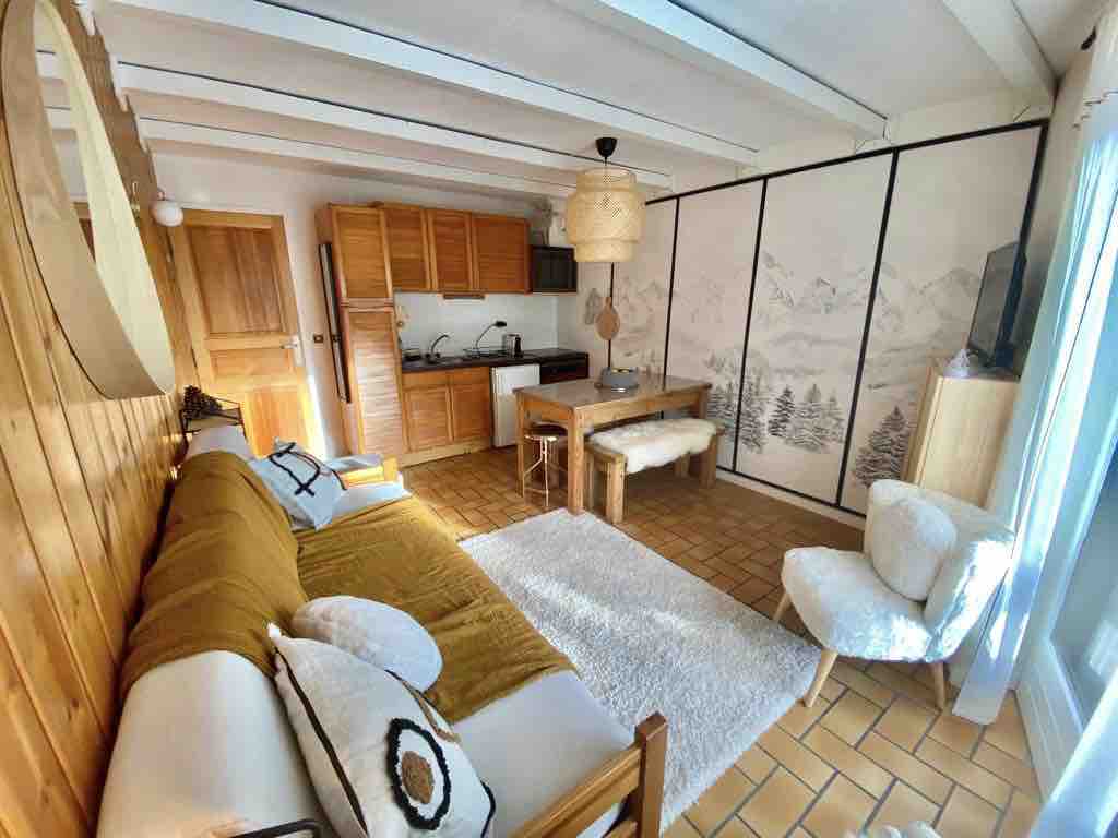A cozy living area features a sofa adorned with cushions and a soft rug underfoot. A wooden table is paired with chairs, while the kitchenette is equipped along one wall. A decorative partition with mountain imagery enhances the space, complemented by natural light coming from the nearby window.