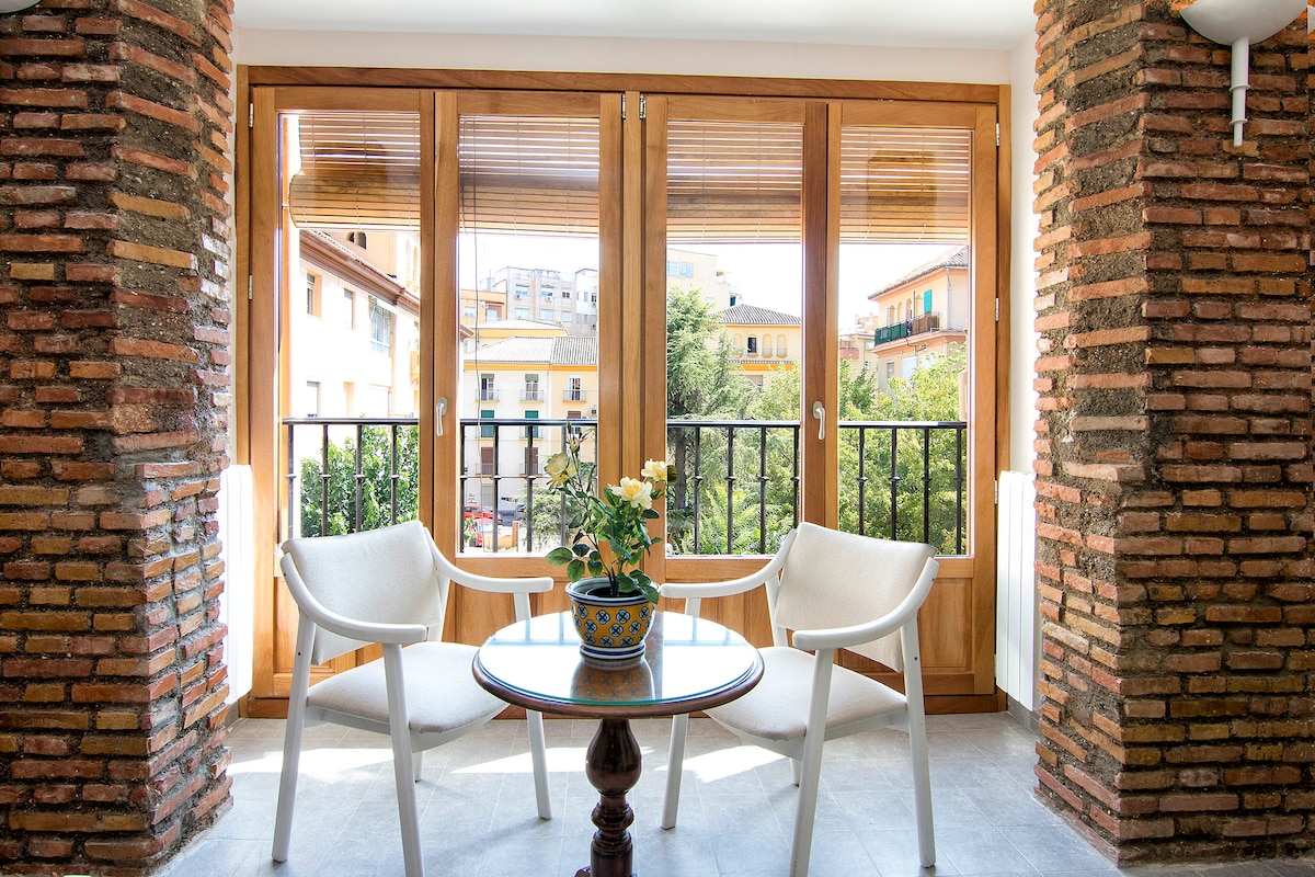 A bright seating area features two white chairs positioned around a round table. Large windows surrounded by rustic brick walls allow natural light to fill the space, offering views of the exterior landscape and nearby buildings.