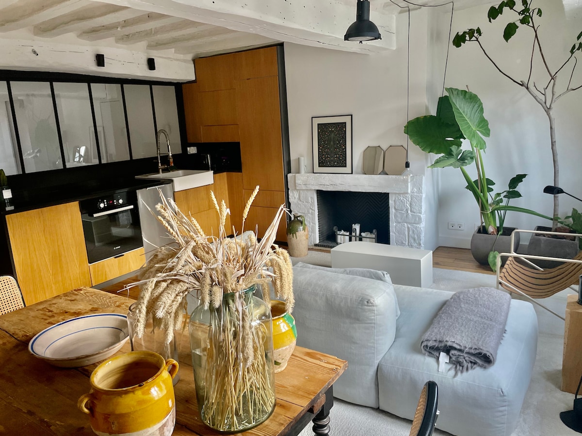 The spacious living area features modern furnishings with a comfortable sofa and a two-tone coffee table. A sleek kitchen is visible in the background, along with a decorative chimney. Houseplants add a touch of greenery, creating a relaxing ambiance.