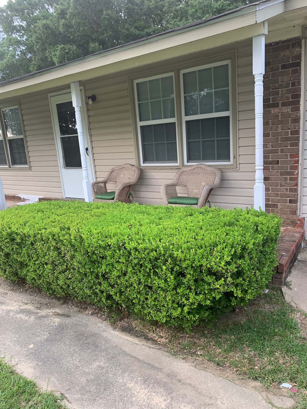 A well-maintained front porch features two wicker chairs positioned beside a neatly trimmed hedge. Large windows allow natural light to illuminate the inviting entryway, creating a calm and welcoming space. The pathway is lined with grass, contributing to the home's overall curb appeal.