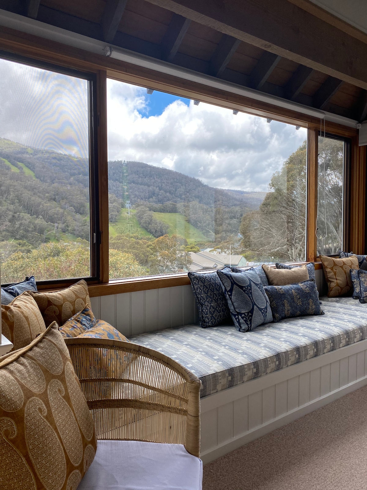 A sunlit seating area features a comfortable bench adorned with various cushions, providing an inviting spot to enjoy views of the surrounding mountains. The large windows bring in natural light while framing the scenic landscape outside.