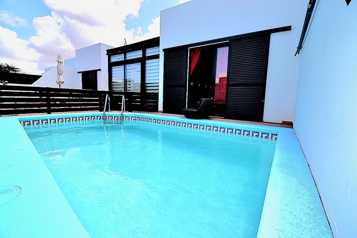 A rectangular pool with clear blue water is framed by a white border. Black sliding doors open to an interior space, revealing large windows and a patio area. Comfortable seating is positioned nearby, with an umbrella providing shade.