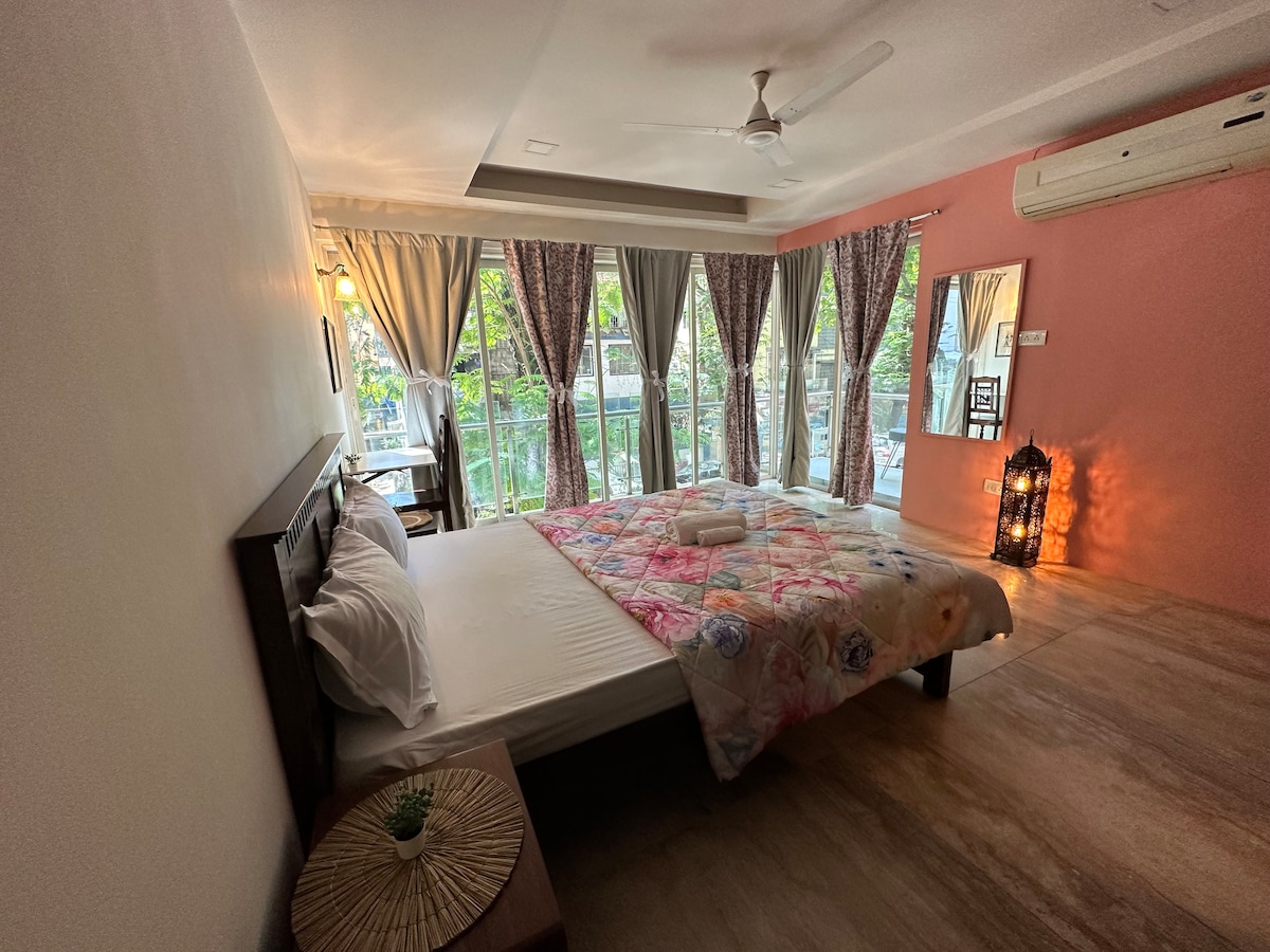A bright sleeping area is furnished with a spacious bed draped in a floral blanket. Large windows provide a view of greenery and natural light, accentuated by elegant curtains. A bedside table and a decorative lantern complement the warm color scheme of the room.