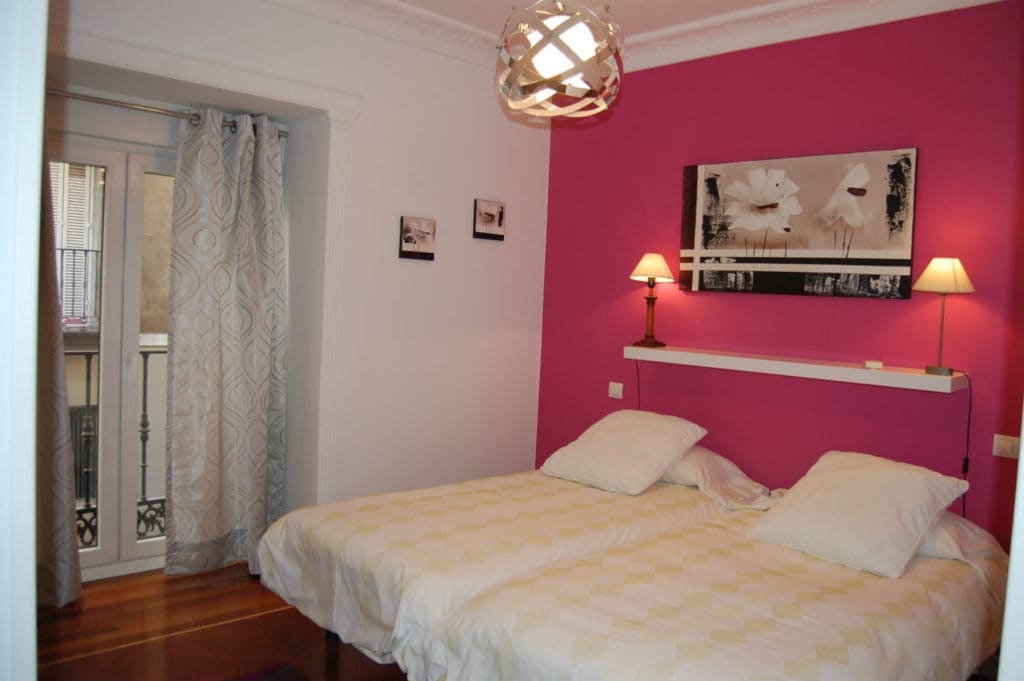 A room with a vibrant pink wall features two individual beds, each with white linens and pillows. A minimalist shelf displays a black-and-white artwork, while lamps on either side add soft lighting. A large window opens to the exterior, complemented by light curtains.