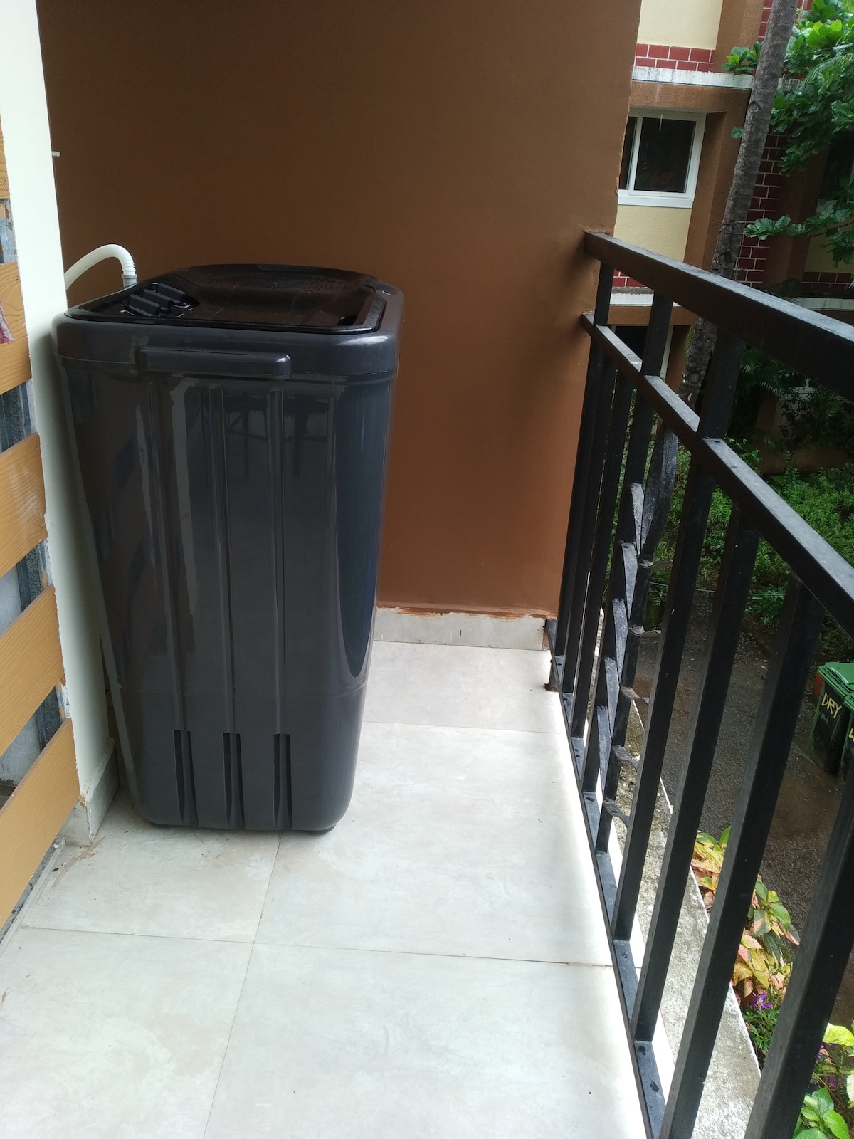 A compact washing machine is positioned on a balcony. The machine is gray and has a top-loading design. The balcony features a tiled floor and black railing, providing a view of the greenery below, enhancing the outdoor living space.