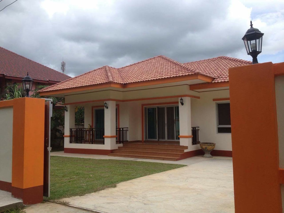 A modern single-story house features a welcoming entrance with a covered porch and steps leading to the front door. The exterior is accented with orange trim and stone elements, surrounded by a well-kept lawn. Lantern-style lights add illumination to the pathway.