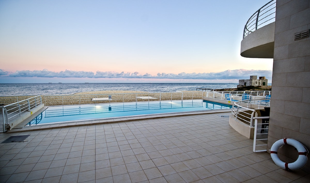 An expansive communal pool area is visible, with a smooth tiled surface extending towards the beach. The calm sea is seen in the background, complemented by a pastel sky at dusk. A safety ring is positioned near the pool's edge, enhancing the tranquil setting.