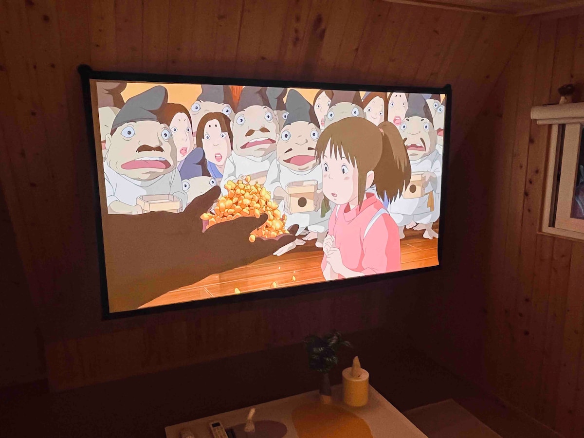 An indoor area features a large projection screen displaying a scene from an animated film. The image captures a girl with a surprised expression, surrounded by characters that appear cartoonish and expressive. A cozy room atmosphere is suggested by the wooden wall paneling.
