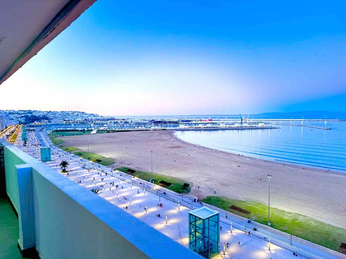 A panoramic view from the balcony reveals a serene beach at dusk, with soft waves lapping at the shore. The beachside promenade is visible, lined with palm trees. The fading light casts a gentle glow over the calm water and the distant marina.