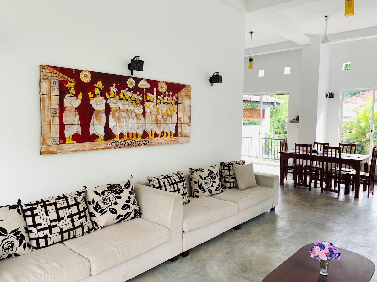 A common living area features a large, light-colored sofa adorned with patterned throw pillows. A vibrant artwork depicting traditional figures hangs on the wall. A dark wooden coffee table sits in front, and a dining area with wooden chairs is visible in the background.