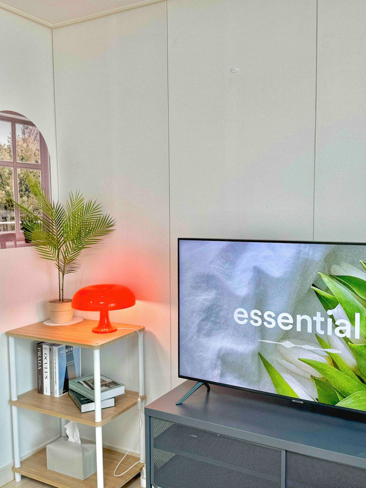 A modern living space features a sleek television displaying the word 'essential.' An orange lamp provides warm light on a bookshelf holding various items, while a green plant adds a touch of nature. A window allows natural light to brighten the area.