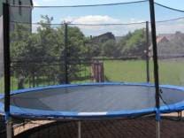 A round trampoline is situated on a grassy surface, enclosed by a safety net. Nearby trees and greenery create a natural backdrop, complementing the outdoor play area. The sky is clear with a few clouds, enhancing the open and inviting space.