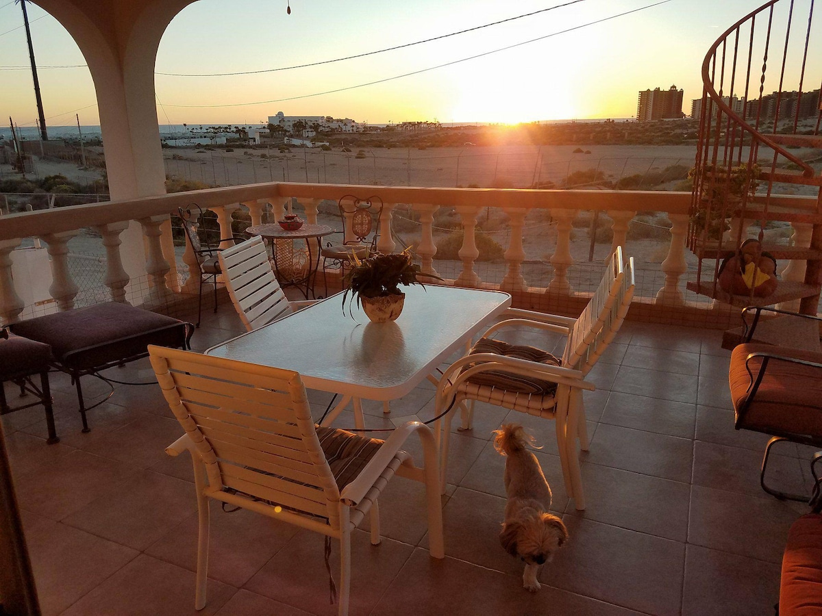 A spacious balcony is depicted with a glass table and white chairs, complemented by a potted plant in the center. The sunset casts a warm glow over the scene, with soft views of distant buildings and the beach beyond. A small dog is seen exploring the area.