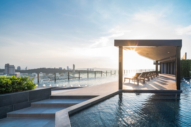 A rooftop pool area is visible, featuring a sleek infinity edge that merges with panoramic views of the city skyline. Sun loungers are positioned under a shaded structure, creating a relaxing space for guests. The scene is bathed in soft evening light, enhancing the tranquil ambiance.