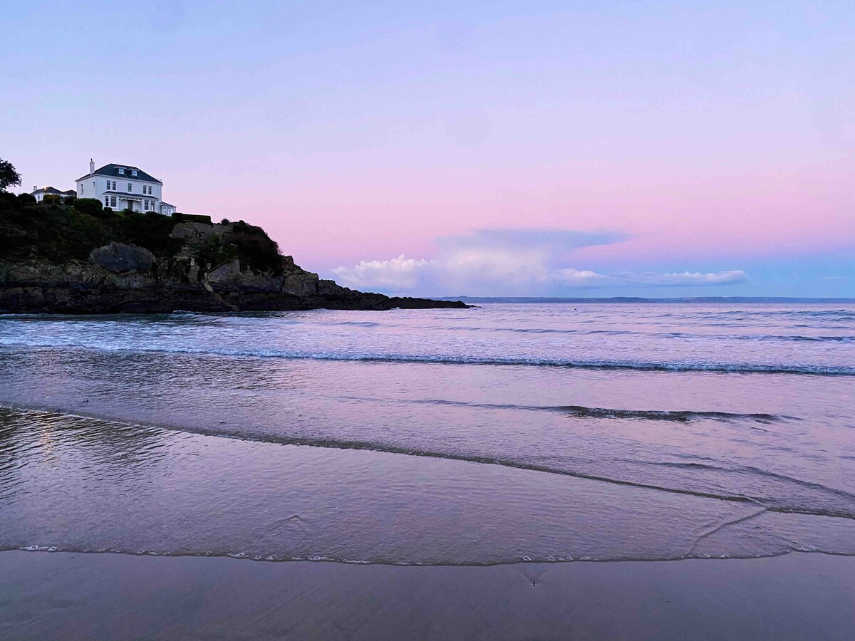 A serene beach scene captures gentle waves lapping at the shore under a pastel sky at dusk. A house is perched on the rocky shoreline, while the horizon fades into soft shades of pink and blue, reflecting a tranquil coastal atmosphere.