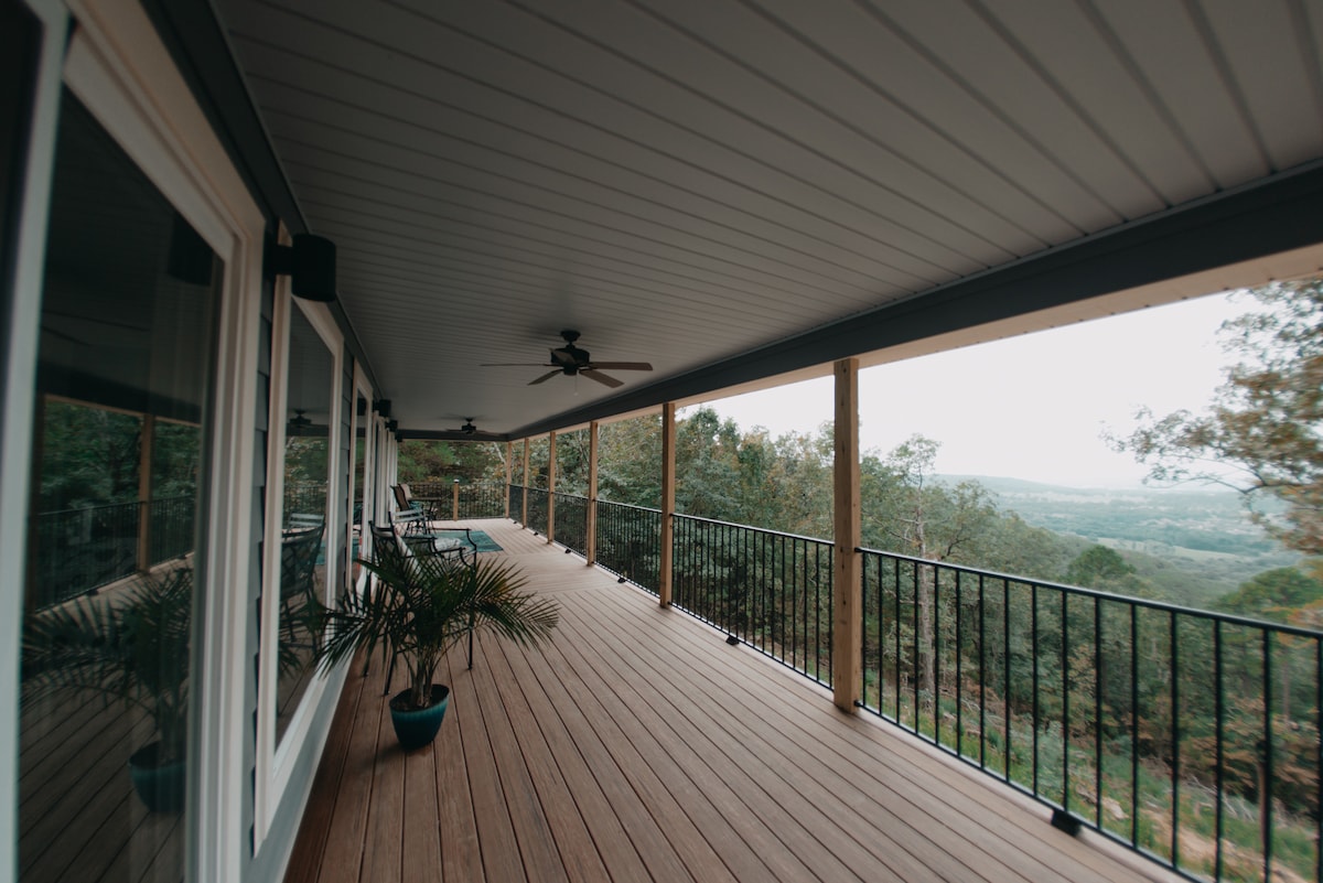 A spacious wooden deck extends along the cottage, featuring several seating arrangements with views of the surrounding nature and distant cityscape. Ceiling fans are installed for comfort, while potted plants add greenery to the area. The railing provides safety without obstructing the view.