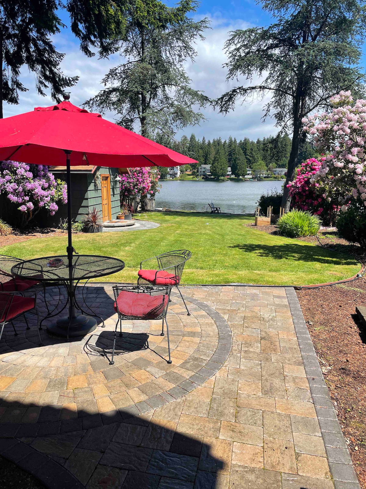A patio area is highlighted by a large red umbrella providing shade over a round table with four chairs. Lush greenery and flowering plants frame the view of the lake in the background, creating a serene outdoor space for relaxation.