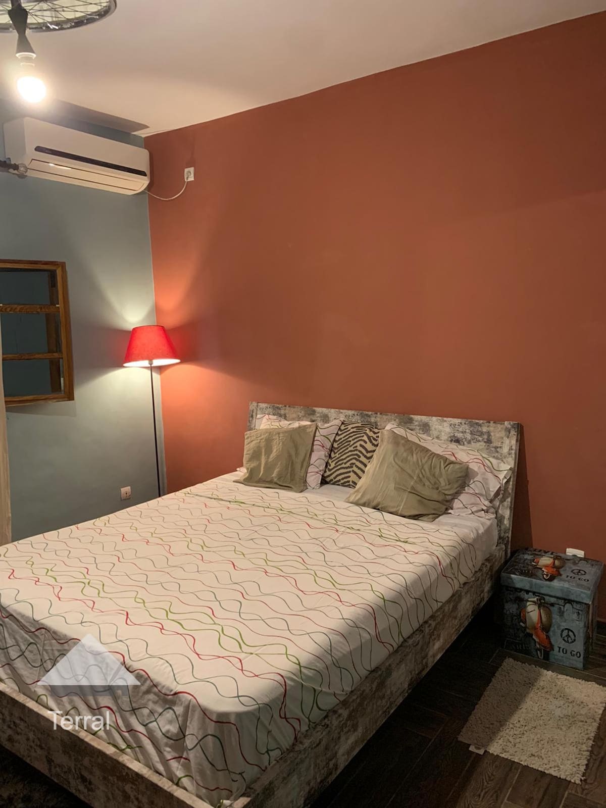 A cozy bedroom features a spacious bed with a patterned coverlet and two decorative pillows. A warm lamp stands beside the bed, casting soft light, while a small accent table is positioned nearby. The walls are painted in calming tones, enhancing the room's inviting feel.