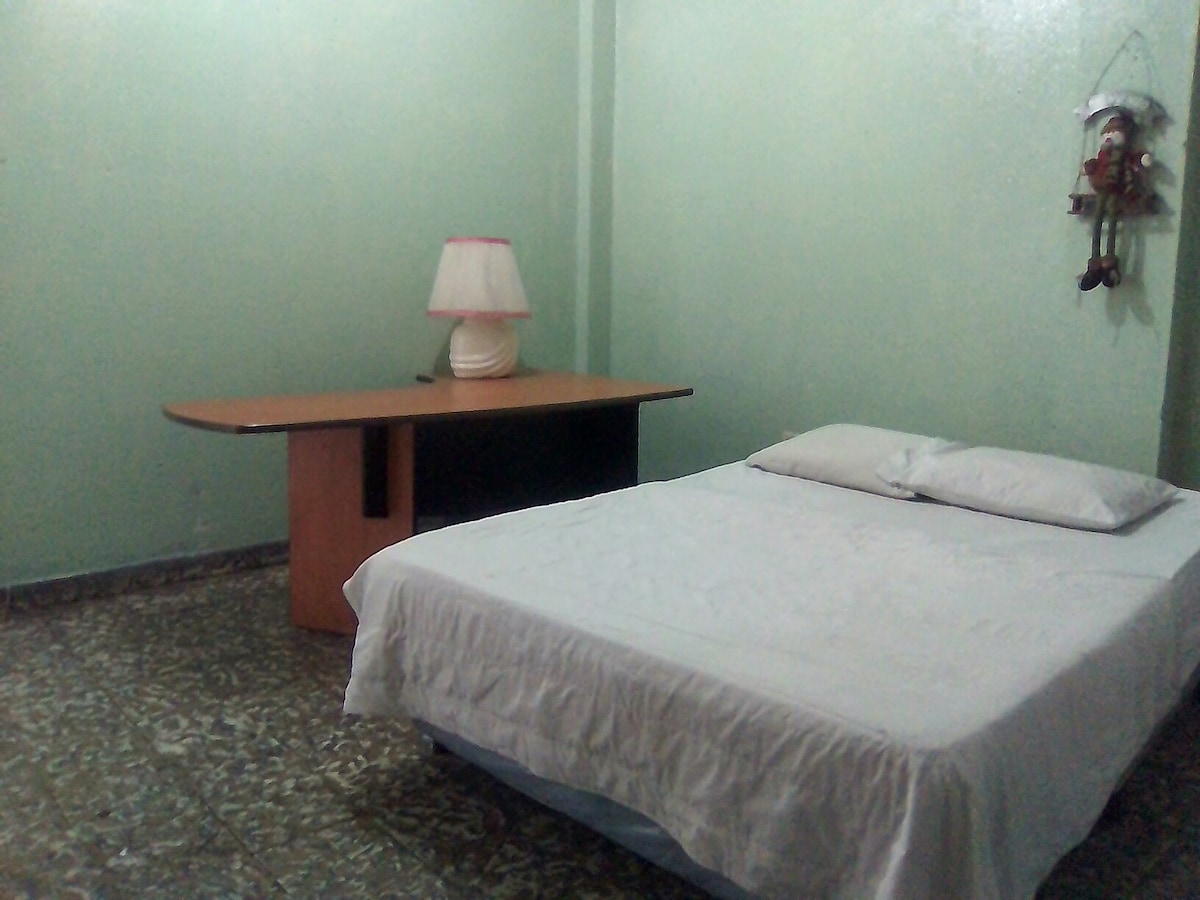 A simple room features a spacious bed with white bedding and a lamp on a nearby table. The green walls and tile floor contribute to the overall fresh ambiance of the space.