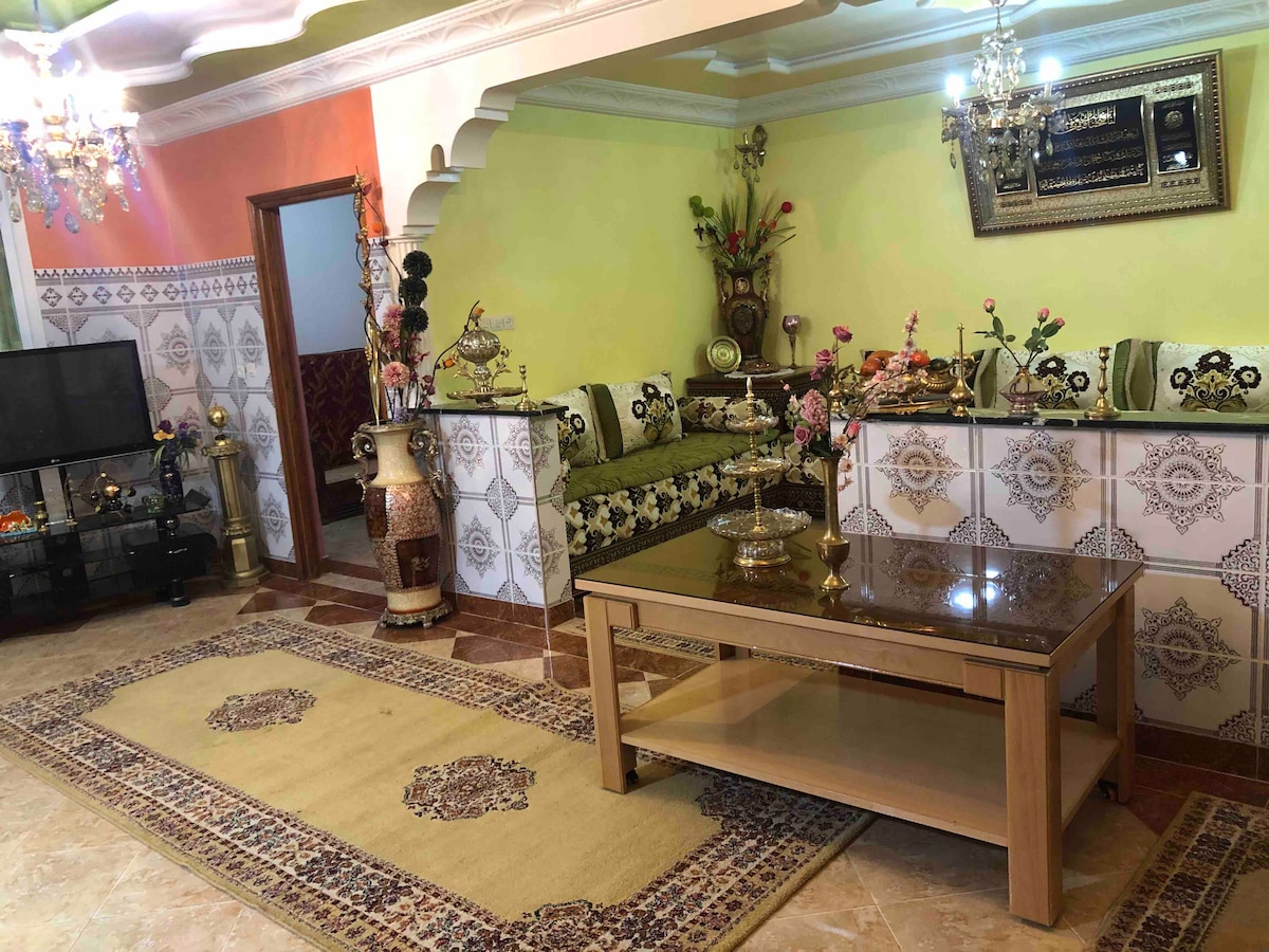 The living room showcases elegant decor with intricate patterns on the sofa and a glass coffee table at its center. Beautiful light fixtures hang from the ceiling, and decorative vases with flowers are placed throughout the space. A television is positioned on a media console against the wall.